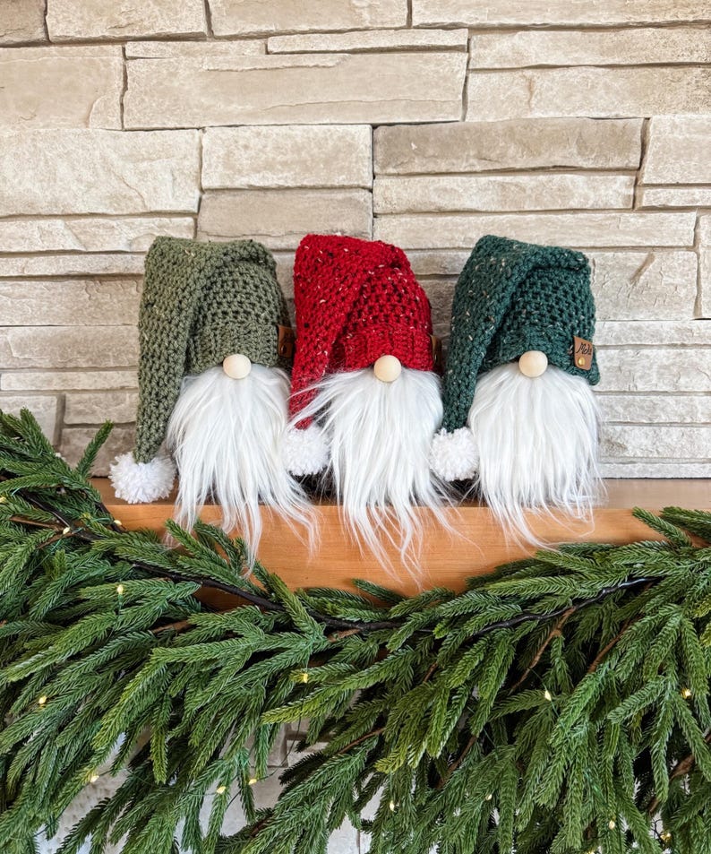 May include: Three gnomes with white beards and knitted hats. The hats are green, red, and green. The gnomes are sitting on a wooden surface with a green garland in front of them.