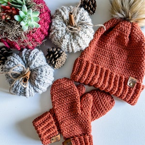 May include: A set of crocheted winter accessories in a burnt orange color. The set includes a beanie with a faux fur pom pom and a pair of mittens. The beanie has a label that reads "Mel's".