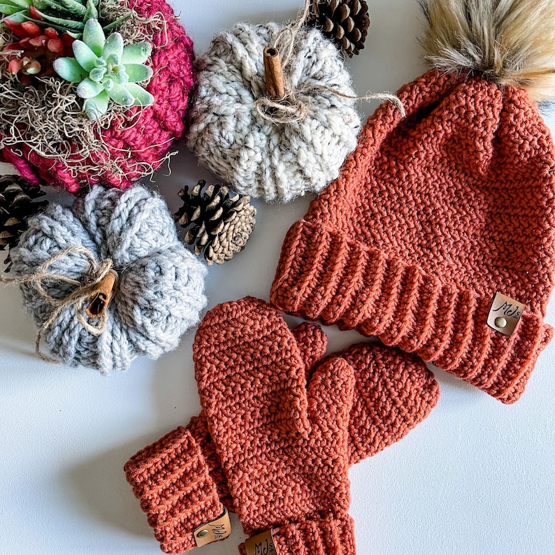May include: A set of crocheted winter accessories in a burnt orange color. The set includes a beanie with a faux fur pom pom and a pair of mittens. The beanie has a label that reads "Mel's".
