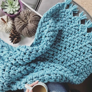 Pu&ograve; includere: Una morbida coperta in crochet blu con frange sui bordi. La coperta &egrave; appoggiata su un vassoio bianco con oggetti decorativi.