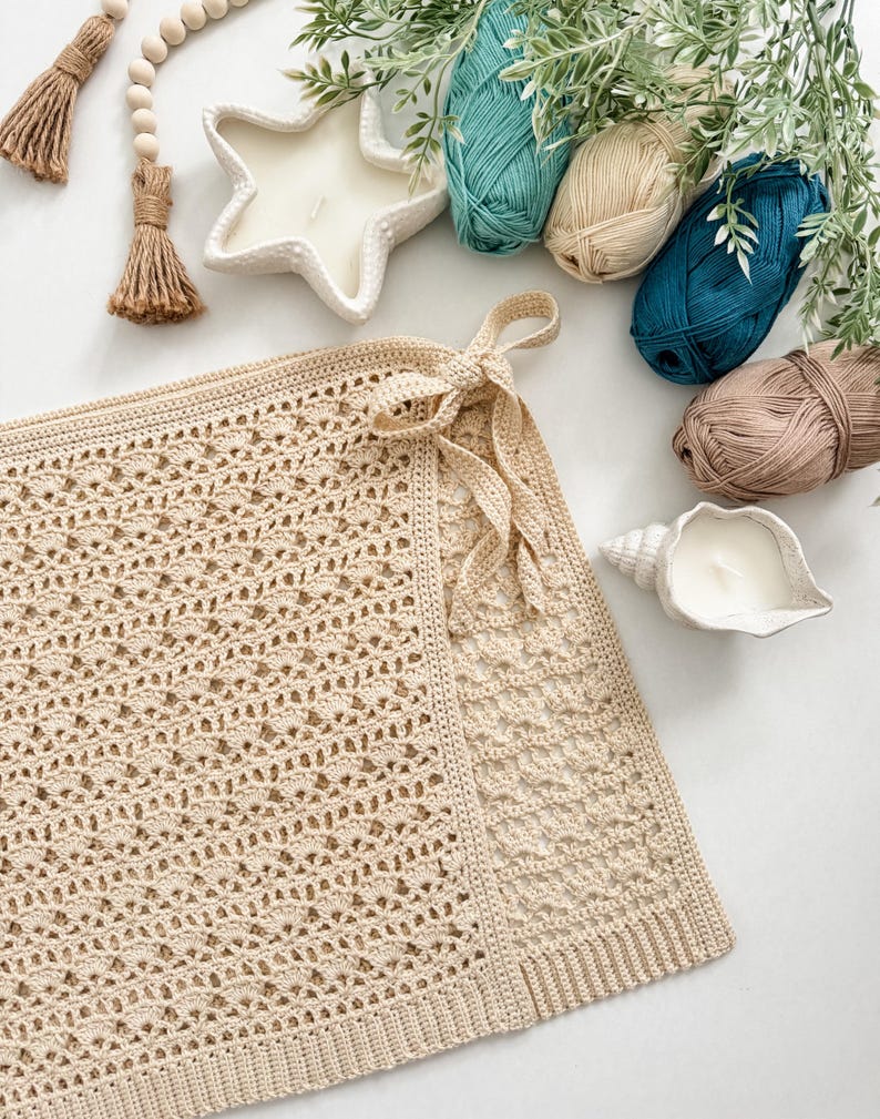 K&ouml;nnte beinhalten: Ein beigefarbener H&auml;kel-Wrap mit Schleifenverschluss, pr&auml;sentiert mit Garnkn&auml;ueln in T&uuml;rkis, Beige und Braun. Dekorative Kerzen in Stern- und Muschelform sind ebenfalls vorhanden, zusammen mit Holzperlen und Gr&uuml;nzeug.