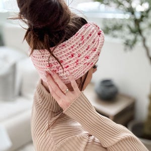 May include: A pink and white crocheted headband with a twisted design. The headband is worn by a person with brown hair pulled back in a bun.