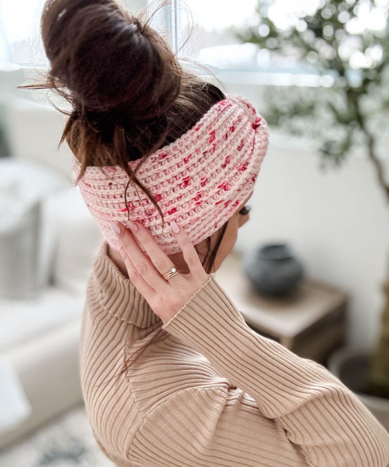 May include: A pink and white crocheted headband with a twisted design. The headband is worn by a person with brown hair pulled back in a bun.