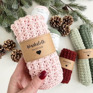 May include: A collection of handmade washcloths and dishcloths in various colors, including pink, burgundy, and green. Each cloth is wrapped with a brown paper band that reads "Washcloth Handmade" or "Dishcloths Handmade" with a heart symbol.