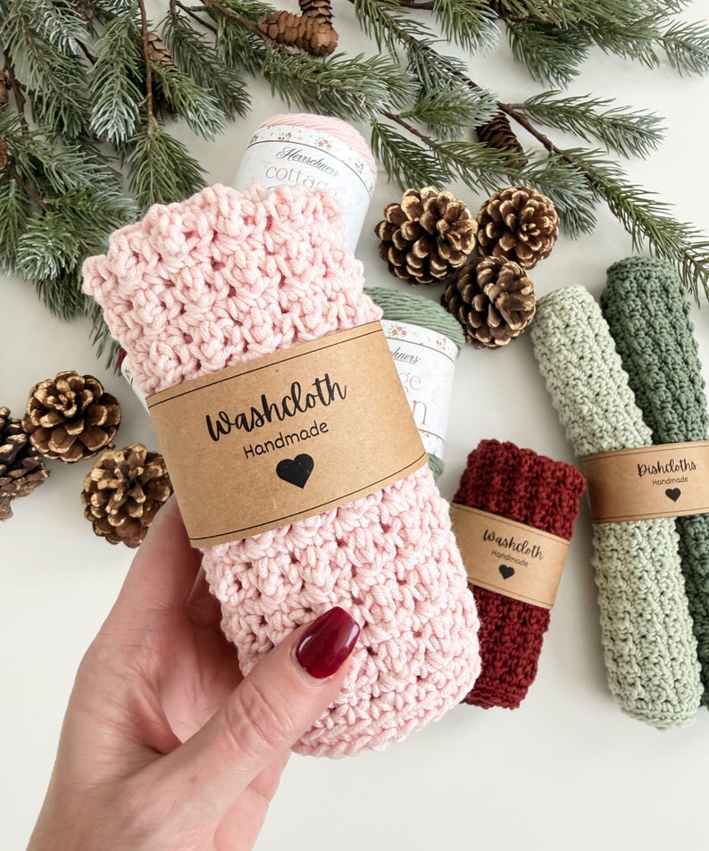 May include: A collection of handmade washcloths and dishcloths in various colors, including pink, burgundy, and green. Each cloth is wrapped with a brown paper band that reads "Washcloth Handmade" or "Dishcloths Handmade" with a heart symbol.