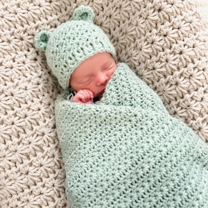 Puede incluir: Una manta de arrullo para beb&eacute; y un gorro a juego tejidos a ganchillo en color verde salvia claro, con dos peque&ntilde;as orejas redondeadas en la parte superior. El conjunto presenta un patr&oacute;n de puntos en forma de estrella en toda la pieza.