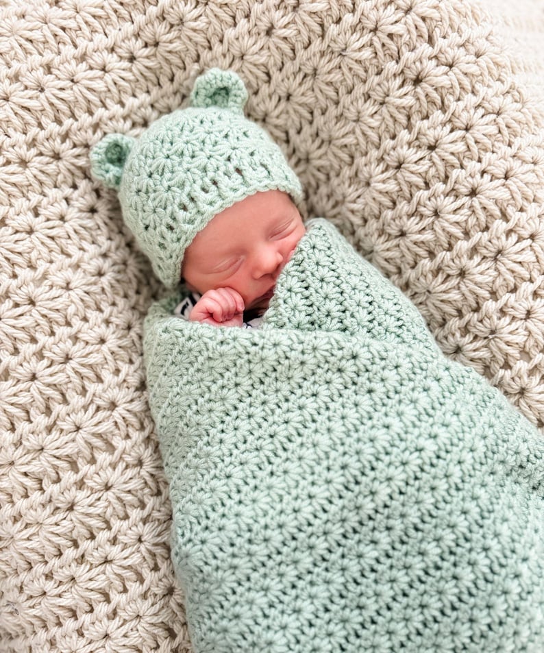 Puede incluir: Una manta de arrullo para beb&eacute; y un gorro a juego tejidos a ganchillo en color verde salvia claro, con dos peque&ntilde;as orejas redondeadas en la parte superior. El conjunto presenta un patr&oacute;n de puntos en forma de estrella en toda la pieza.