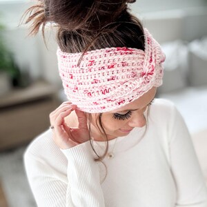 May include: A pink and red crocheted headband with a knotted design. The headband is worn by a person with brown hair.