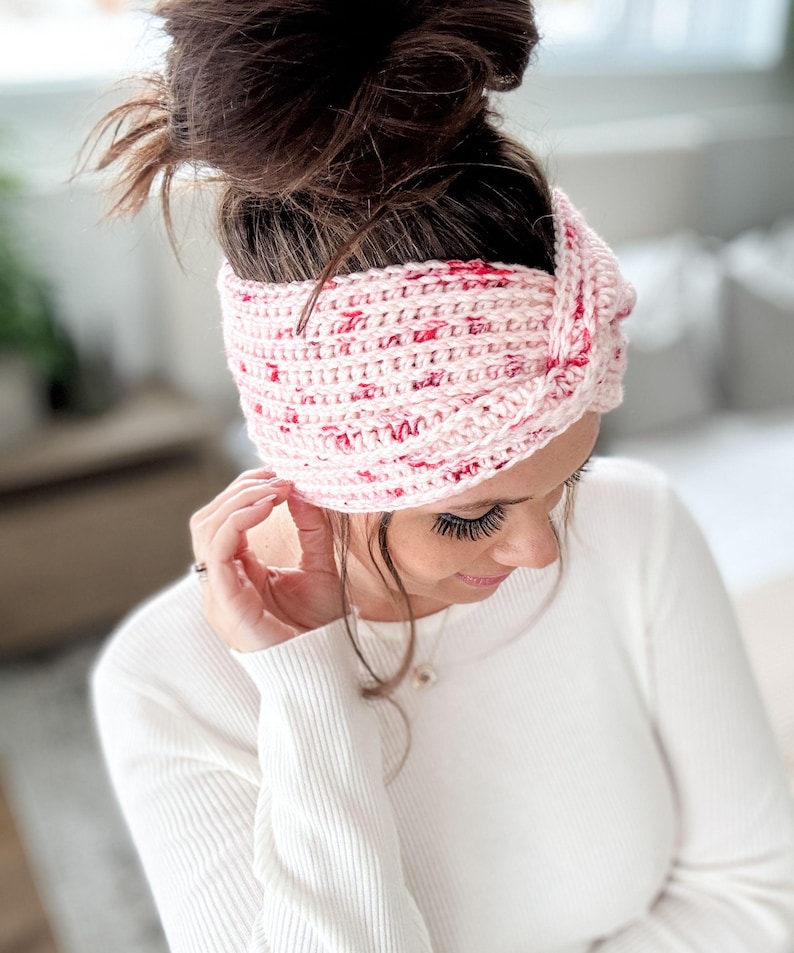 May include: A pink and red crocheted headband with a knotted design. The headband is worn by a person with brown hair.