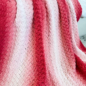 May include: A handmade crochet blanket in shades of red and pink. The blanket features a textured stitch pattern and a gradient color effect, transitioning from deep red to light pink. The soft, cozy blanket is perfect for adding warmth and style to any space.