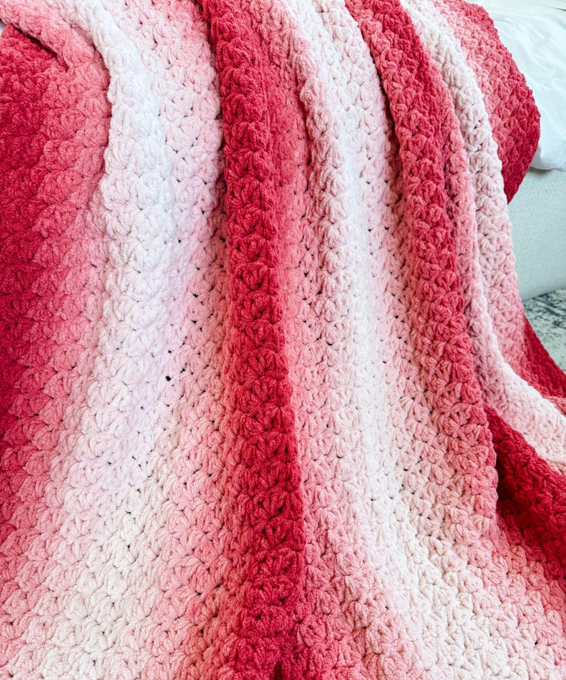 May include: A handmade crochet blanket in shades of red and pink. The blanket features a textured stitch pattern and a gradient color effect, transitioning from deep red to light pink. The soft, cozy blanket is perfect for adding warmth and style to any space.