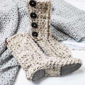 May include: A pair of beige crocheted slipper boots with black buttons and a brown leather strap. The boots have a suede sole.