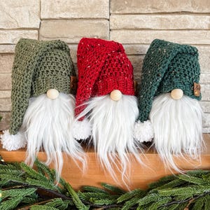 May include: Three crocheted gnomes with white fur beards and pom-poms on their hats. The gnomes are wearing green, red, and green hats.