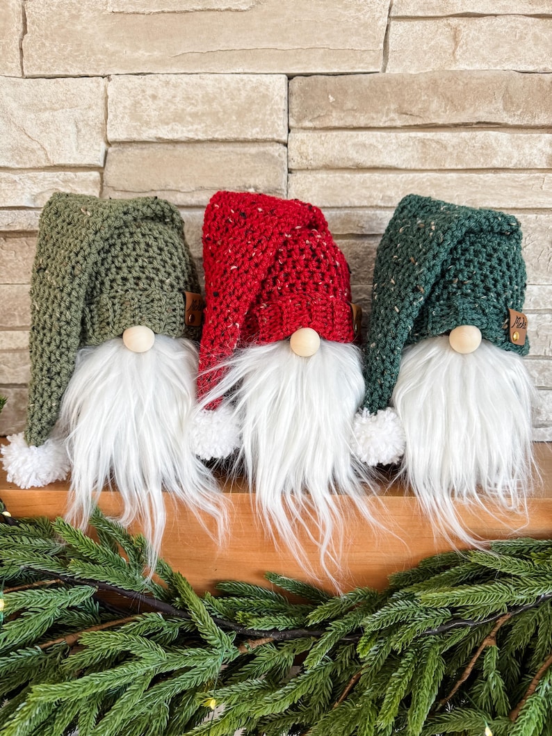 May include: Three crocheted gnomes with white fur beards and pom-poms on their hats. The gnomes are wearing green, red, and green hats.