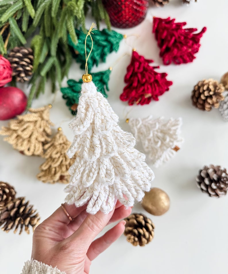 K&ouml;nnte beinhalten: Handgefertigte Weihnachtsbaumschmuck in Wei&szlig;, Gold, Gr&uuml;n und Rot. Die Ornamente, mit goldenen Akzenten, sind als Weihnachtsb&auml;ume geformt. Tannenzapfen und andere festliche Dekorationen sind ebenfalls zu sehen.