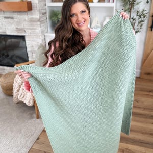Puede incluir: Una manta rectangular de ganchillo en color verde salvia que presenta un patr&oacute;n de puntos texturizados consistente en toda la superficie. La manta tiene un borde definido con una estructura de puntos ligeramente distinta en los extremos.