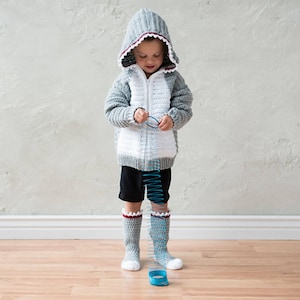 May include: A child wearing a gray and white crocheted shark hoodie with a red zipper. The hoodie has a white crocheted shark fin on the back. The child is holding a blue slinky toy.