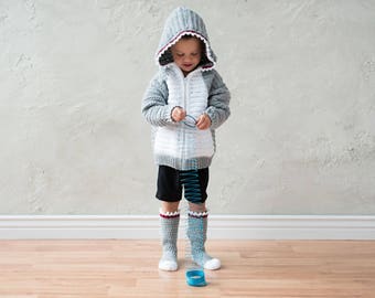 Puede incluir: Un niño lleva una sudadera con capucha de tiburón de crochet gris y blanco con una cremallera roja. La sudadera tiene una aleta de tiburón de crochet blanco en la espalda. El niño sostiene un juguete slinky azul.