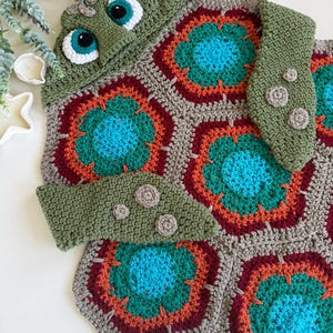 May include: A crochet hooded blanket designed to resemble a sea turtle. The blanket features a shell made of hexagonal motifs in shades of teal, orange, and dark red, bordered by beige yarn. The hood includes large eyes with teal irises.