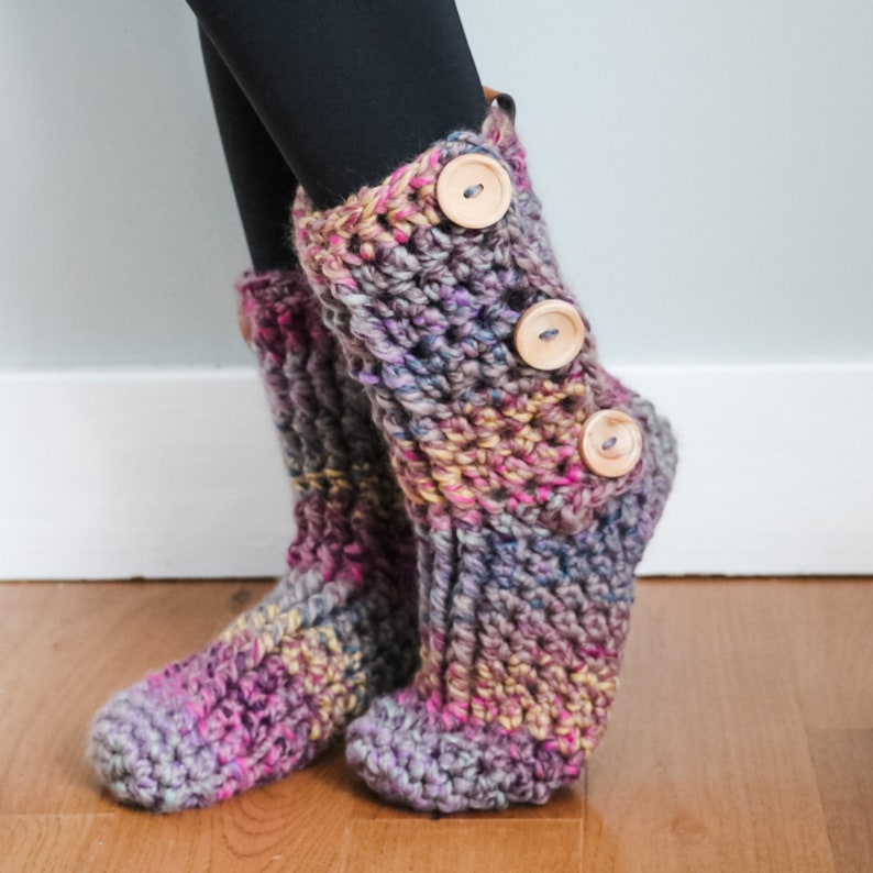 May include: A pair of crocheted boot cuffs made with multicoloured yarn in shades of pink, purple and yellow. The cuffs have three wooden buttons on the side.