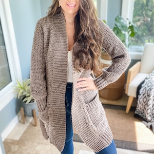 Op de afbeelding: Een lange, bruine cardigan met een shawlkraag en zakken. De cardigan is gehaakt en heeft een losse pasvorm.