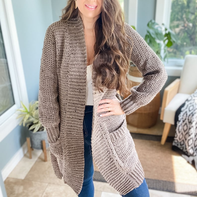 Op de afbeelding: Een lange, bruine cardigan met een shawlkraag en zakken. De cardigan is gehaakt en heeft een losse pasvorm.