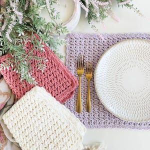 May include: A set of three rectangular crochet placemats in dusty rose, cream, and light purple. The placemats feature a textured, open-weave pattern with scalloped edges. A white ceramic plate with a sunburst pattern and two gold forks are placed on the purple placemat.