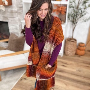 May include: A crocheted shawl in shades of orange, brown, and cream with fringe and pockets. The shawl is draped over the shoulders. The woman is wearing a purple long-sleeve shirt and blue jeans.