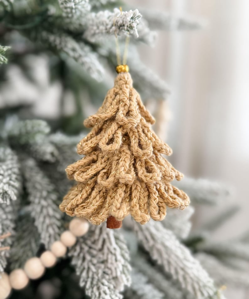 K&ouml;nnte beinhalten: Handgefertigter, goldfarbener Weihnachtsbaumschmuck. Der Baum besteht aus gestricktem Garn mit strukturiertem Design und einem kleinen Holzstamm. Er h&auml;ngt an einem goldenen Faden und wird an einem beschneiten Weihnachtsbaum pr&auml;sentiert.