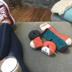 May include: A close-up of a pair of knitted socks in orange, teal, and white with a ball of orange yarn and a ball of dark gray yarn. The socks are on a light gray surface.