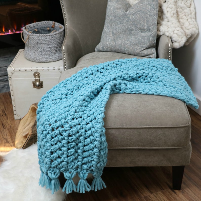 Pu&ograve; includere: Una coperta di uncinetto blu chiaro con frange a nappe drappeggiata su una poltrona grigia. La coperta &egrave; realizzata con un filato spesso e ha un motivo a punto testurizzato.