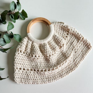 May include: A cream-colored crocheted bag with a wooden handle. The bag has a rounded shape and a delicate, open-weave design.