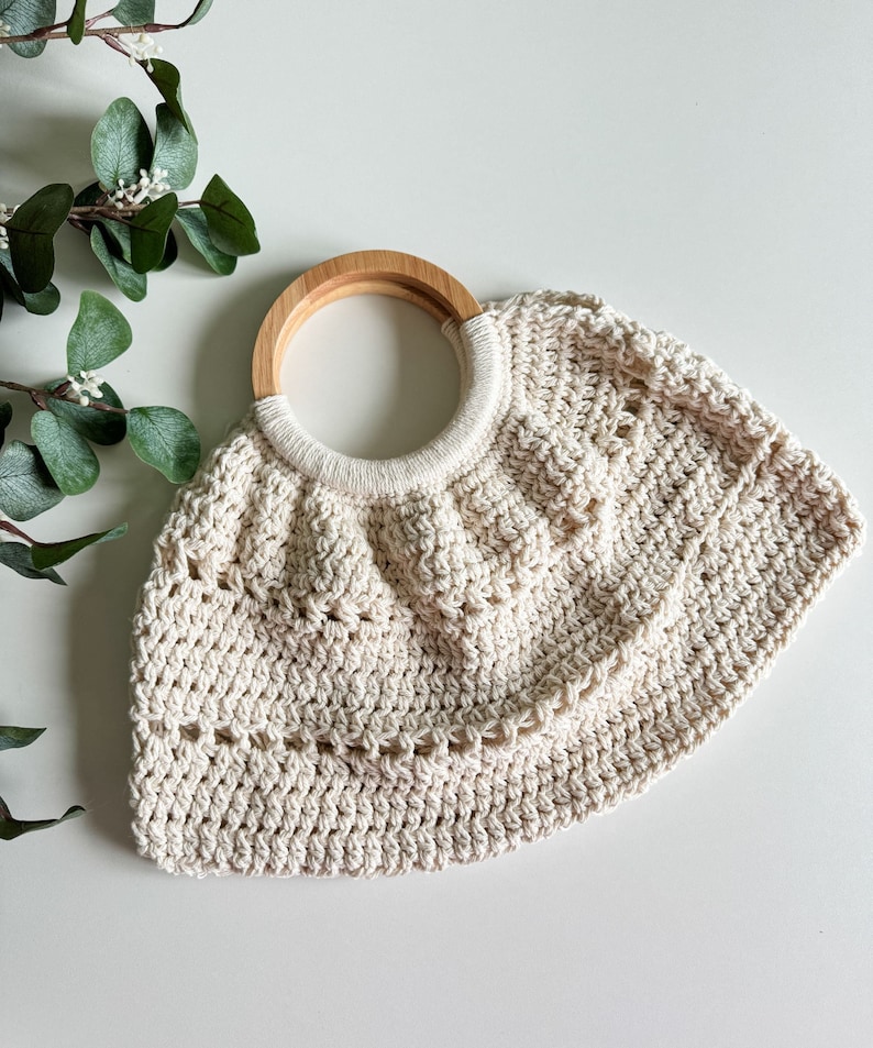 May include: A cream-colored crocheted bag with a wooden handle. The bag has a rounded shape and a delicate, open-weave design.