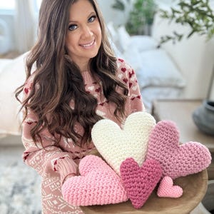 Pu&ograve; includere: Una collezione di cinque cuscini a forma di cuore all'uncinetto in tonalit&agrave; di rosa e bianco. I cuscini sono disposti su un vassoio di legno.