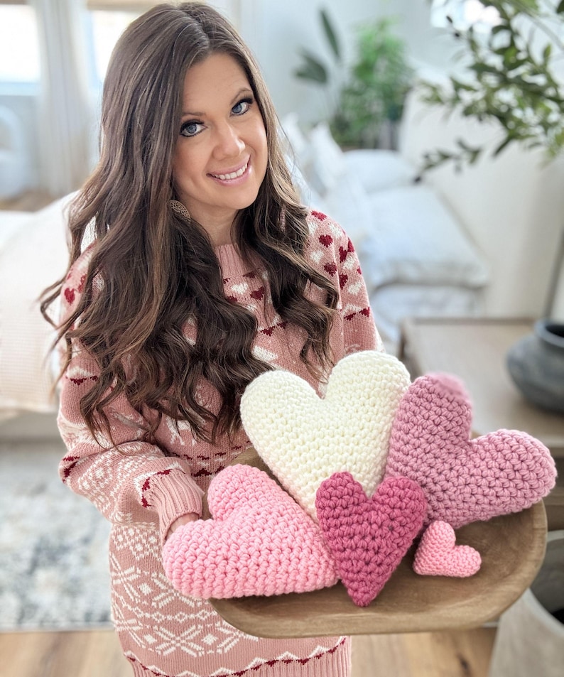 Pu&ograve; includere: Una collezione di cinque cuscini a forma di cuore all'uncinetto in tonalit&agrave; di rosa e bianco. I cuscini sono disposti su un vassoio di legno.