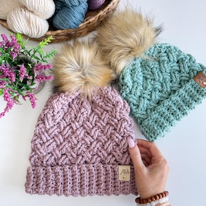 May include: Two crocheted hats with faux fur pom poms. The hats are in a light pink and a light green colour. Both hats have a leather tag with the text "Mel's" on them.