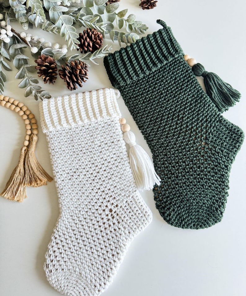 May include: Two handmade Christmas stockings, one white and one dark green, are displayed. The white stocking has a cream-coloured cuff and a white tassel. The green stocking has a matching green tassel. Both stockings have wooden bead accents.