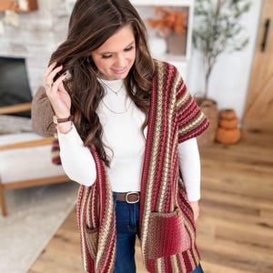 May include: A striped crochet cardigan in shades of red, brown, and beige. The open-front cardigan features two front pockets and a brown belt. The sleeves are short, and the garment is worn over a white long-sleeved shirt and blue jeans.