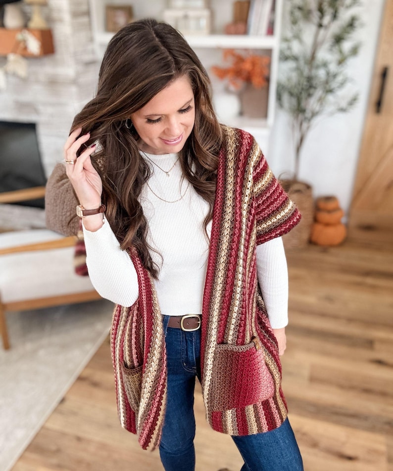 May include: A striped crochet cardigan in shades of red, brown, and beige. The open-front cardigan features two front pockets and a brown belt. The sleeves are short, and the garment is worn over a white long-sleeved shirt and blue jeans.
