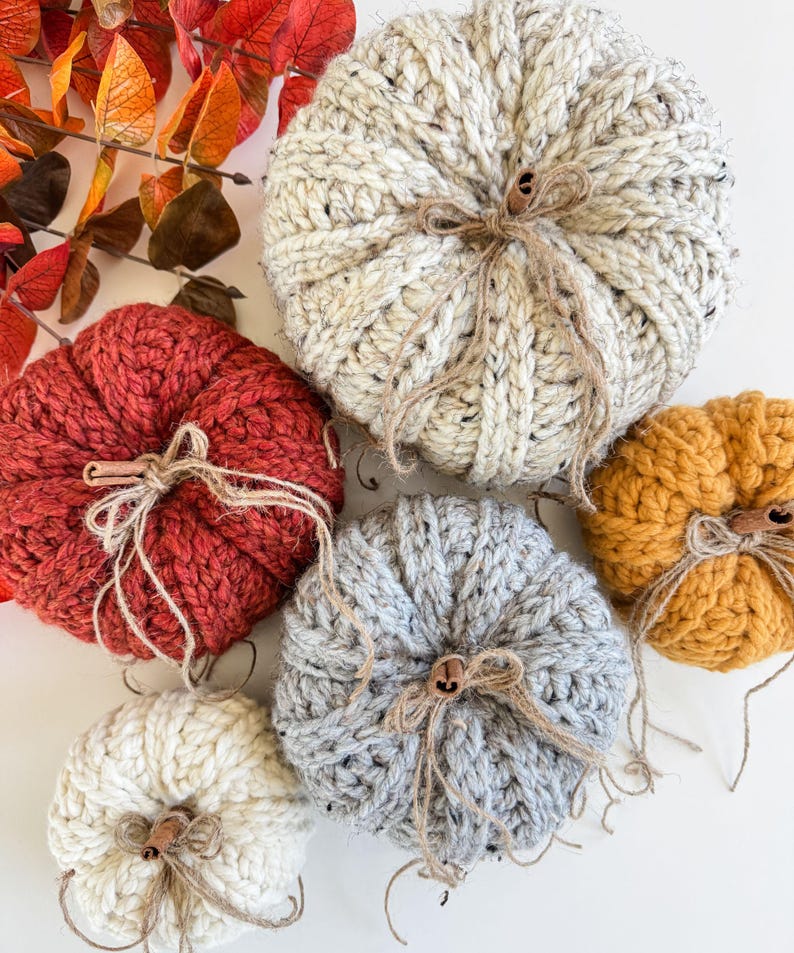 K&ouml;nnte beinhalten: Eine Sammlung handgestrickter K&uuml;rbisse in verschiedenen Farben, darunter Creme, Rot, Grau und Orange. Jeder K&uuml;rbis hat einen Bindfadenstiel und ein kleines Holzknopfdetail, ideal f&uuml;r die Herbstdekoration.