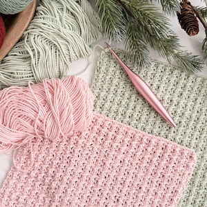 May include: A craft scene featuring pink and green yarn, crochet hooks, and completed crochet squares. The yarn is arranged with a wooden tray, and the crochet squares are in progress. The scene is decorated with pine branches and pine cones.
