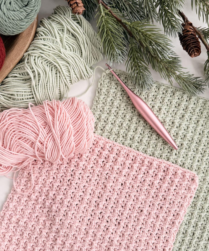 May include: A craft scene featuring pink and green yarn, crochet hooks, and completed crochet squares. The yarn is arranged with a wooden tray, and the crochet squares are in progress. The scene is decorated with pine branches and pine cones.