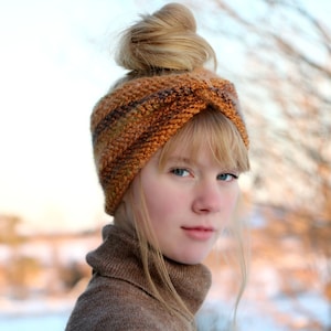 May include: A woman wearing a brown and orange knitted headband with a twisted design. The headband is worn over her hair, which is styled in a bun. She is wearing a brown turtleneck sweater.
