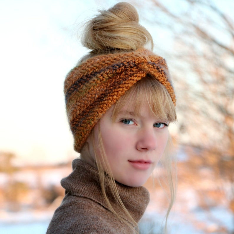 May include: A woman wearing a brown and orange knitted headband with a twisted design. The headband is worn over her hair, which is styled in a bun. She is wearing a brown turtleneck sweater.