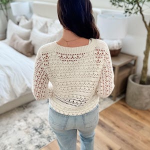 K&ouml;nnte beinhalten: Cremefarbener H&auml;kelpullover mit langen &Auml;rmeln. Der Pullover hat ein offenes, spitzenartiges Design und einen runden Ausschnitt. Die Person tr&auml;gt hellblaue Jeans. Der Hintergrund umfasst ein Bett, einen Beistelltisch und eine Topfpflanze.