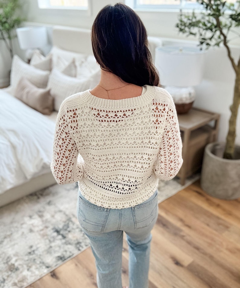 K&ouml;nnte beinhalten: Cremefarbener H&auml;kelpullover mit langen &Auml;rmeln. Der Pullover hat ein offenes, spitzenartiges Design und einen runden Ausschnitt. Die Person tr&auml;gt hellblaue Jeans. Der Hintergrund umfasst ein Bett, einen Beistelltisch und eine Topfpflanze.