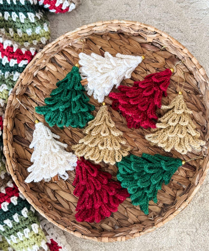 K&ouml;nnte beinhalten: Handgefertigte Weihnachtsbaum-Ornamente aus H&auml;kelarbeit in einem Weidenkorb. Die Ornamente sind in verschiedenen Farben gehalten, darunter Gr&uuml;n, Rot, Wei&szlig; und Gold. Jeder Baum hat eine goldene Aufh&auml;ngeschlaufe. Der Korb steht auf einer neutralen Oberfl&auml;che.