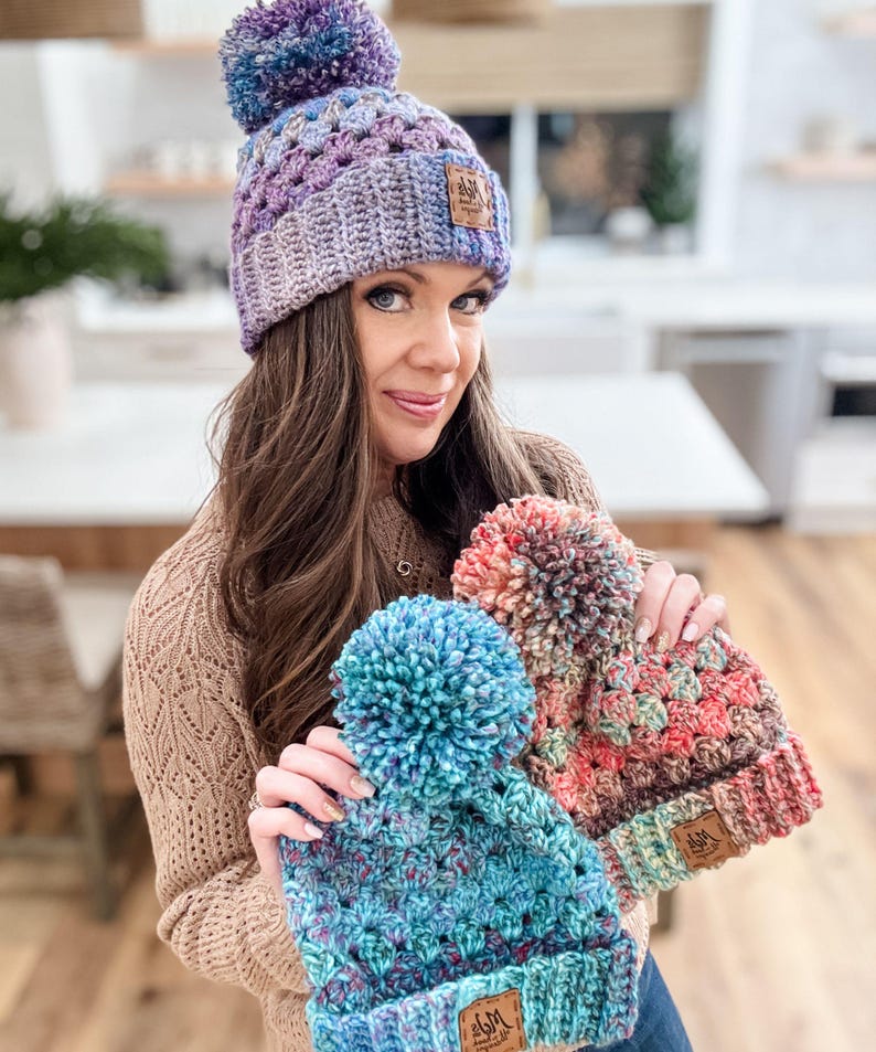 May include: Three crocheted hats with pom-poms in shades of purple, blue, and red. Each hat has a brown leather patch with the text "JM Handmade" on it.