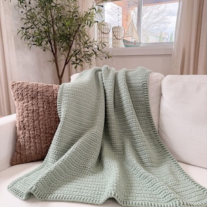 Puede incluir: Una manta de ganchillo color verde salvia con un patr&oacute;n de puntos texturizado y repetitivo, adem&aacute;s de un borde definido. La manta est&aacute; colocada sobre un sof&aacute; blanco, mostrando su color suave y uniforme y su construcci&oacute;n artesanal consistente.