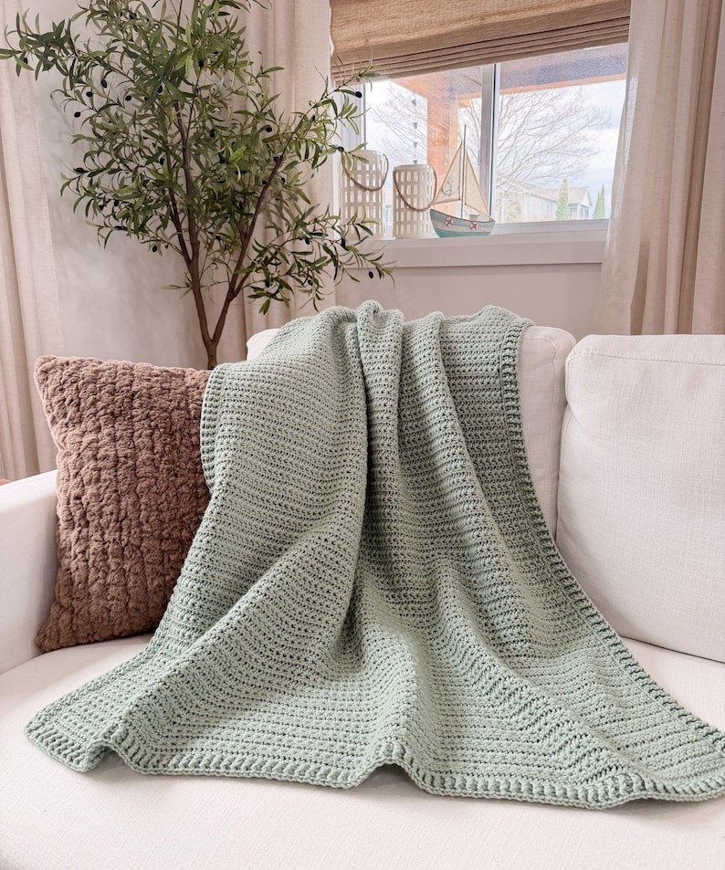 Puede incluir: Una manta de ganchillo color verde salvia con un patr&oacute;n de puntos texturizado y repetitivo, adem&aacute;s de un borde definido. La manta est&aacute; colocada sobre un sof&aacute; blanco, mostrando su color suave y uniforme y su construcci&oacute;n artesanal consistente.